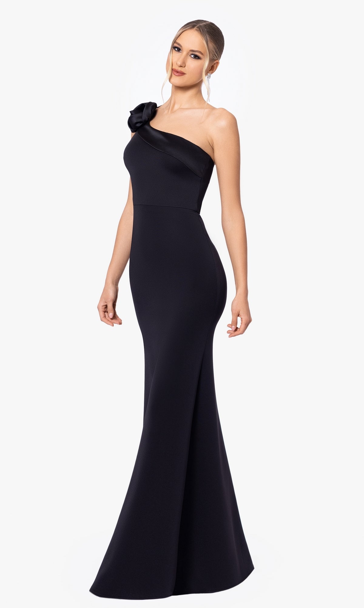 A woman models the Long Black Prom Dress: Blondie Nites 5047BN, a fitted, floor-length, one-shoulder gown with floral strap detail, highlighting its classic elegance against a plain white background.
